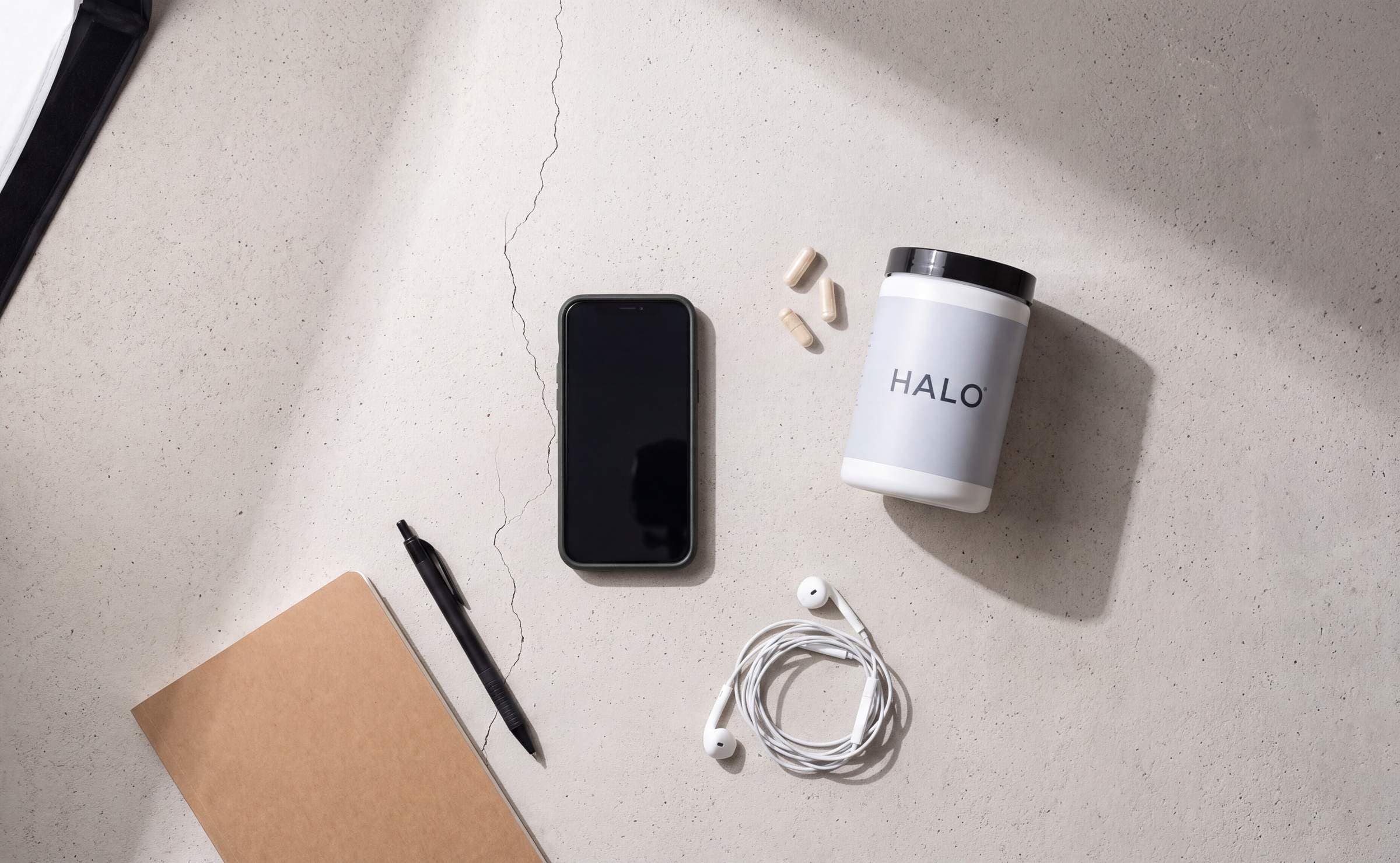 Overhead flat lay of a HALO supplement jar, a phone, a notebook and earbuds on pale concrete