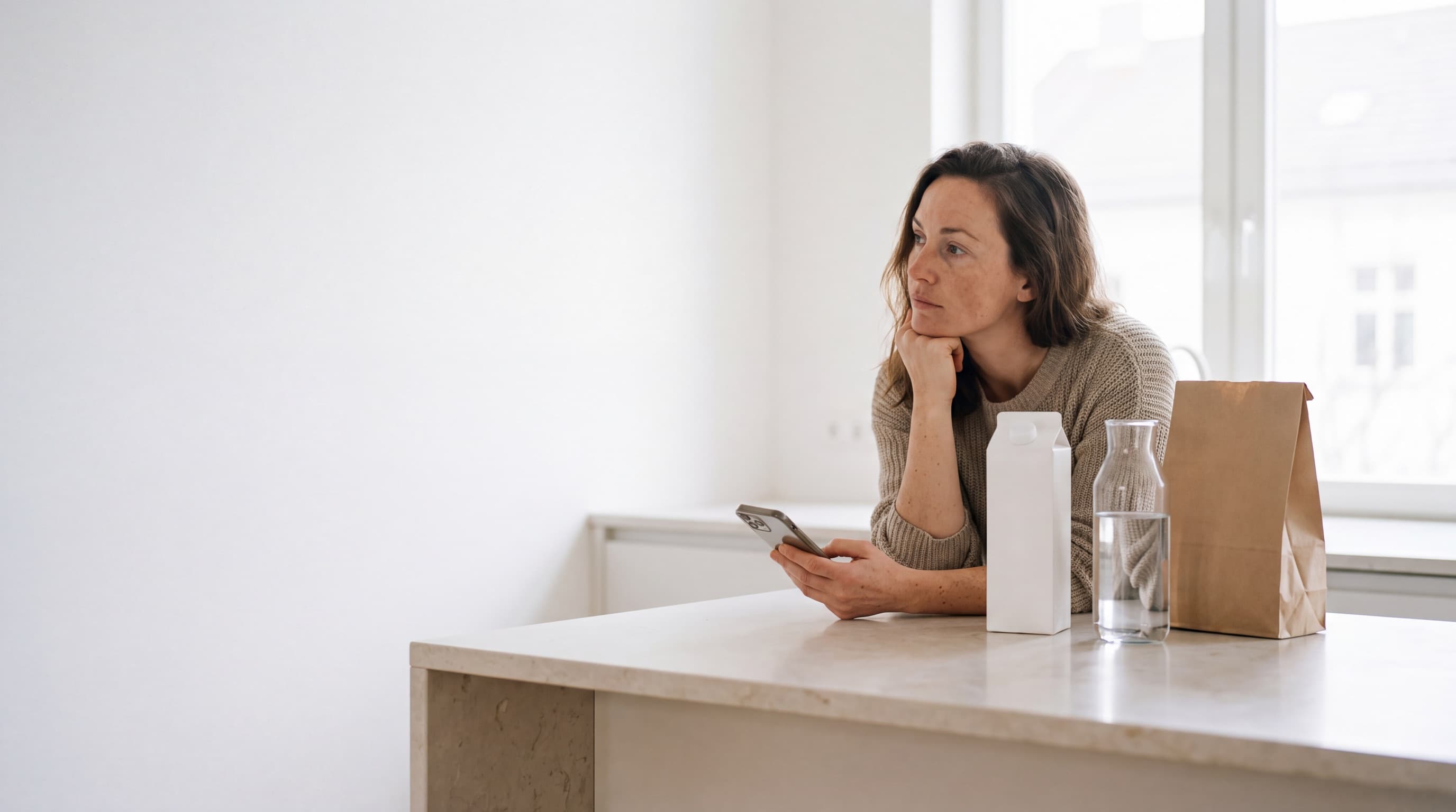 Woman at a kitchen island with her phone in hand, thinking, beside a plain carton, a glass carafe and a kraft bag