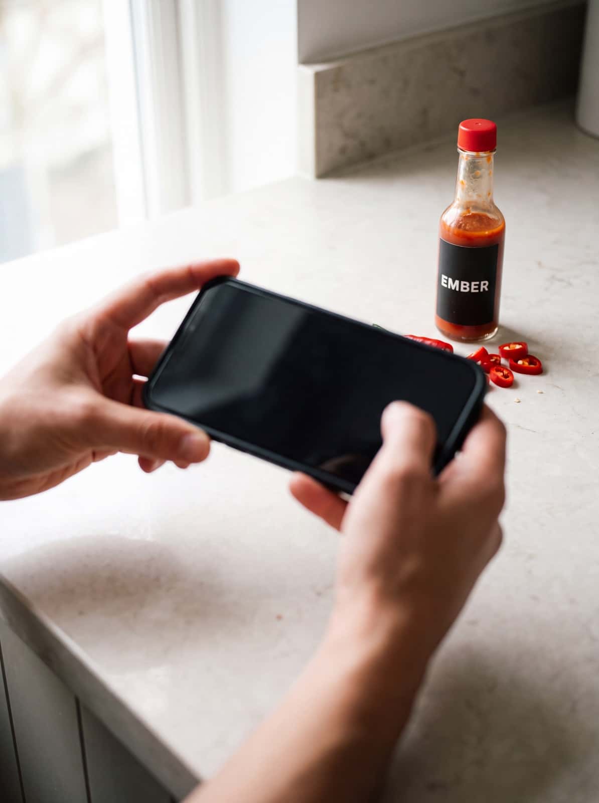 Hands holding a phone up to an EMBER hot sauce bottle and red chillies on a counter