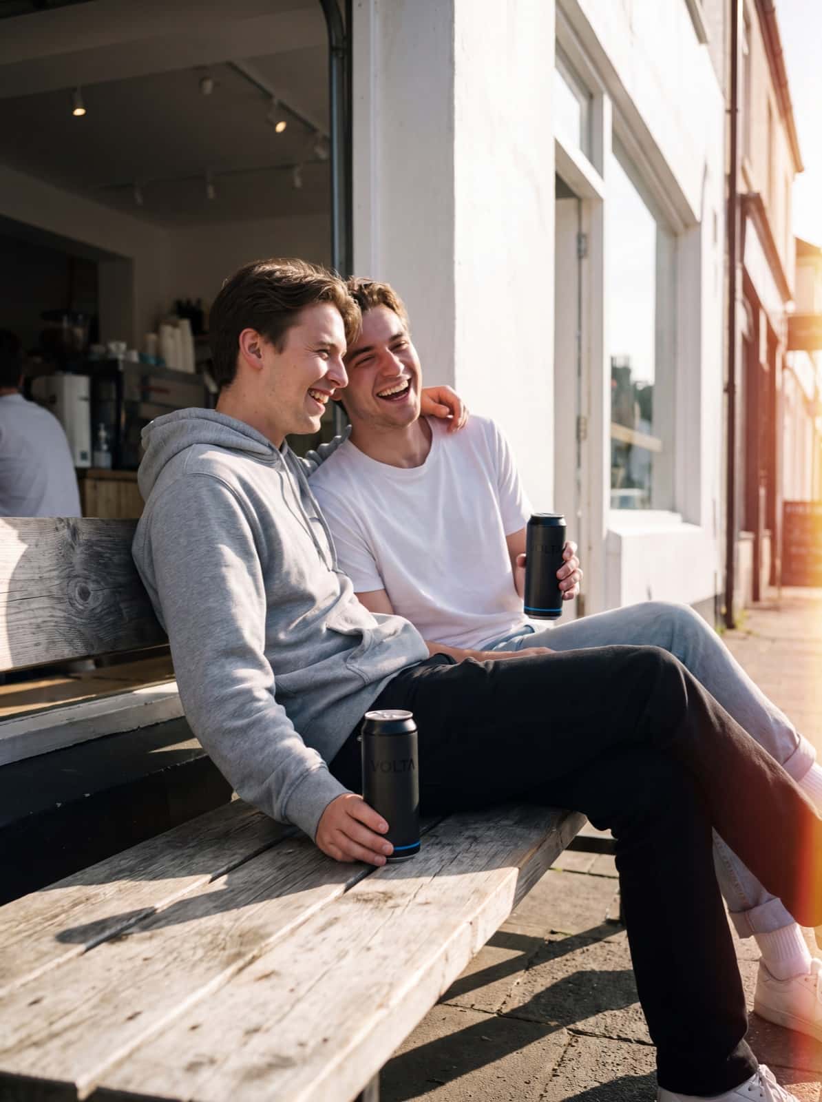 Two friends laughing on a cafe bench, each holding a VOLTA can
