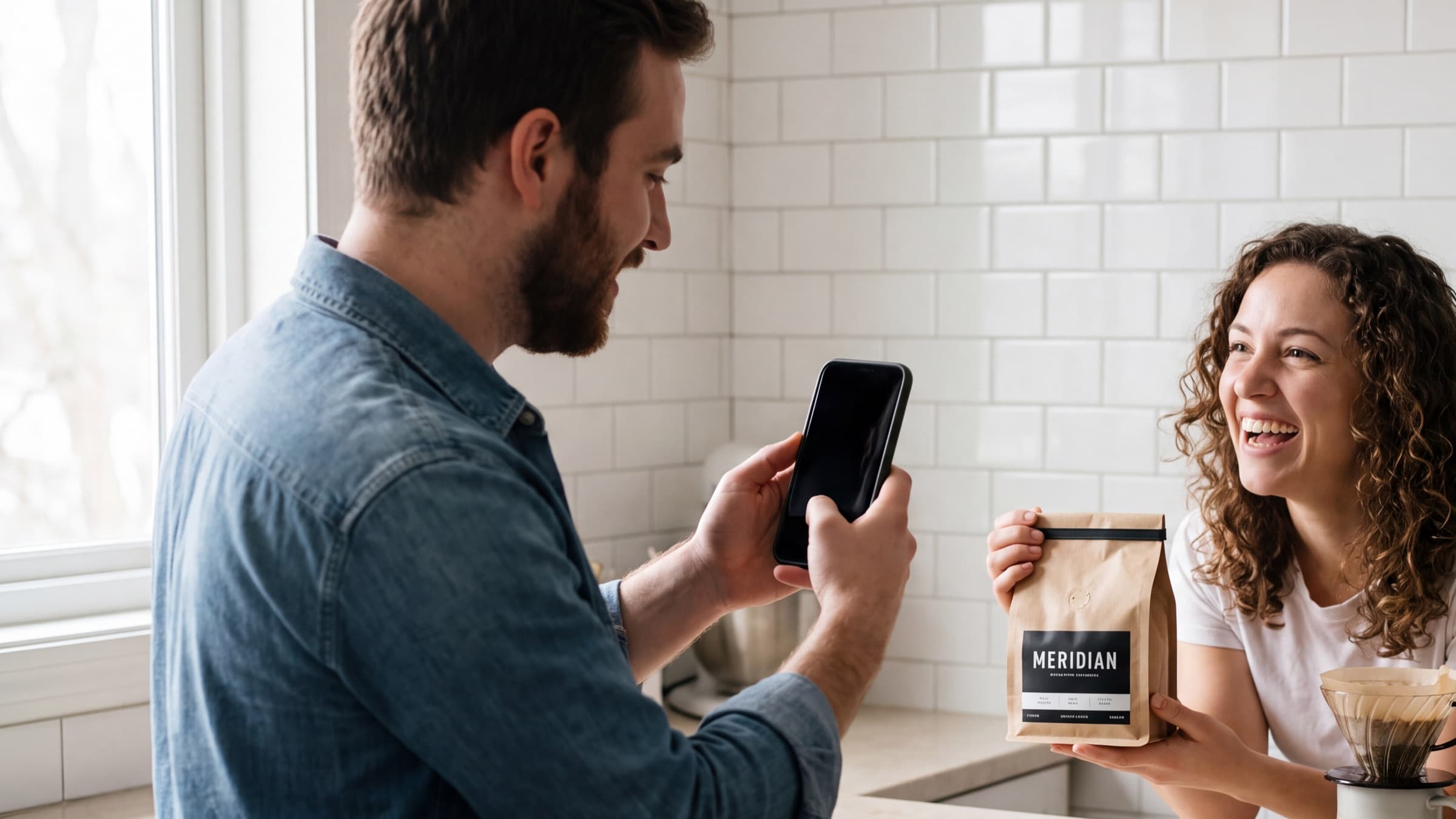 Man filming a friend on his phone as she laughs with a MERIDIAN coffee bag at a kitchen counter